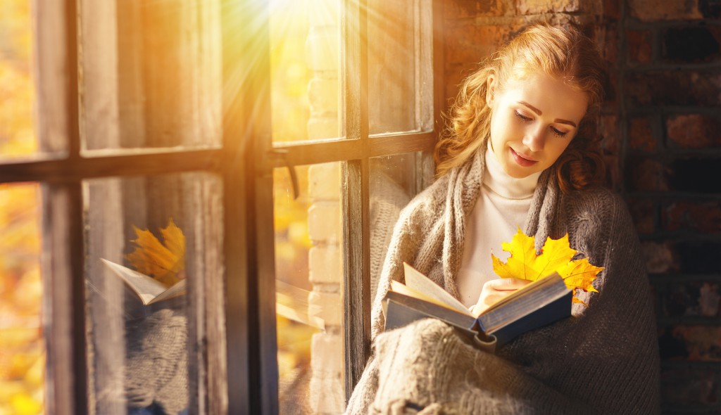 Happy young woman reading book by window in fall